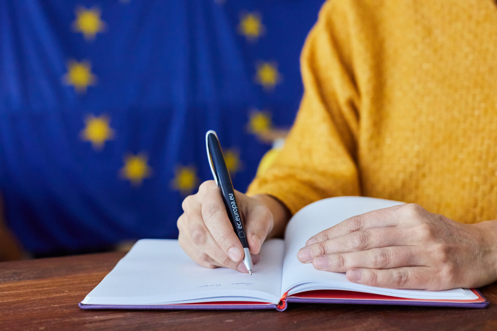 Person håller i EU-penna och skriver i ett block med EU-flaggan i bakgrunden.
