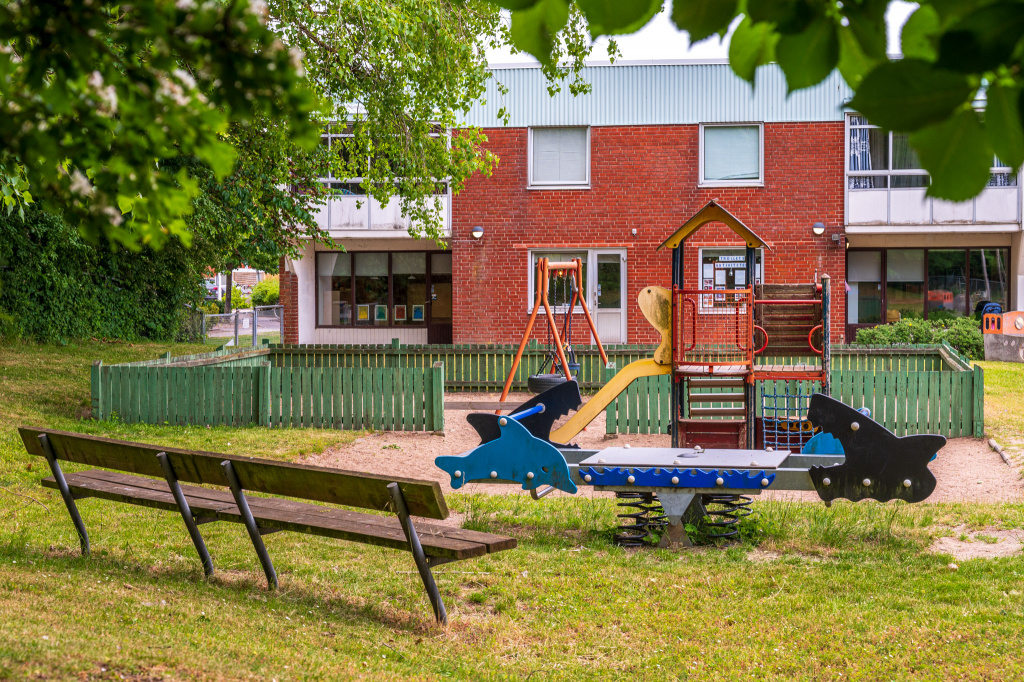 I förgrunden en parkbänk framför en lekplats, omgiven av gräs. I bakgrunden ett tvåplanshus i rött tegel.