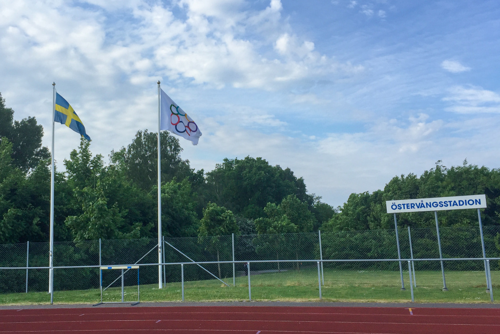 Två flaggstänger, en svensk flagga och en OS-flagga. Skylt med texten Östervångstadion.