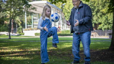 Barn och äldre man trixar med fotboll i park.