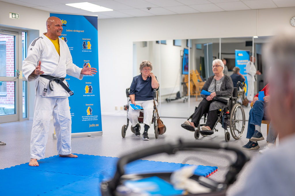 Man i judodräkt pratar inför besökare i rullstol och rollator.
