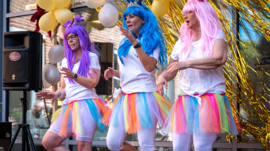 Tre personer står på en scen, utklädda med färgglada peruker.